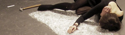 A photo of a performance in which a pale person in a black long-sleeved leotard and tights lays down and caresses what appears to be a rectangle of broken glass. A hammer sits on the floor at their feet.