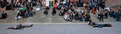 A photo of a broad stair to access a building. Groups of people sit on the steps and two people lie on the ground, seemingly unmoving, in the foreground