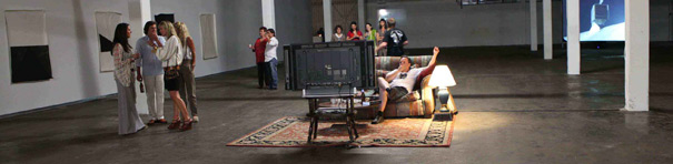 A photo of a dimly lit white-walled gallery space. Some two dimensional items are hung on the walls and a video plays at the back. The center of the image is brightened by a lamp sitting on a area rug with a couch and television. A person sits on the couch apparently watching television while clusters of people are arrayed behind this scene throughout the columned space.