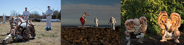 First image: outside on a sunny day, in the middle ground on a lawn, two men in hazmat suits stand about four feet apart, holding a large white ring, behind a complex sculpture of an imaginary creature on the ground. In the second image, on a wide swath of rocky ground against a cloudy skyscape, the two men stand near a third very tall sculptural figure. The third image is a close-up of two imaginary sculptural creatures with extremely large ears, beneath leafy bushes.