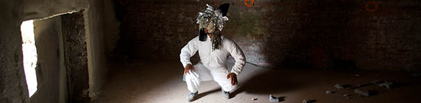 This wide rectangular image shows a crouching figure front and center, wearing a white long-sleeved outfit. A silvery mask covers their face and head. An opening on the far left of the scene lets slivers of bright light into the darkened concrete interior space.