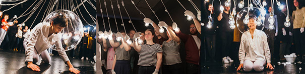 "Three images, all with bright light bulbs dangling from above. In the first, a white-clad performer is kneeling on stage, hands pressed to the floor. Their face is seen behind a large clear bubble. Behind in a semicircle, a dozen or so performers each hold a long wire connected to a light bulb. The second image is a close-up of people who appear to be audience members holding the lightbulbs. In the third image, the performer kneels with the audience behind, the same bubble over their head, and a bucket and props on the stage behind. "