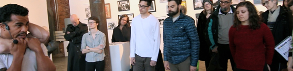 The photo depicts a mic'd swarthy man with short curly dark hair in the foreground—he is being choked from behind by an offscreen figure, both of his hands grasping the choker's forearms. Two observers face the scene, one looking and the other a bald man with head bowed. The image also shows 9 people in an indoor art gallery with framed pictures on the wall and a white pedestal, watching something in front of them—their facial expressions all appear quite concerned.