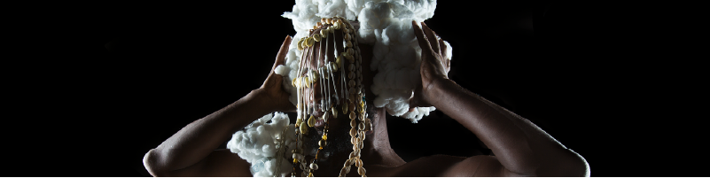 A photo of a Black person wearing a headdress made of a beaded scrim that covers their face and helmet of cotton balls. They hold their hands to their head and the photo is cropped at their elbows. The background is black.