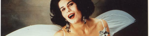 In a grey room, a white woman with dark hair and white wings looks into the camera. She is seen from the shoulders up.