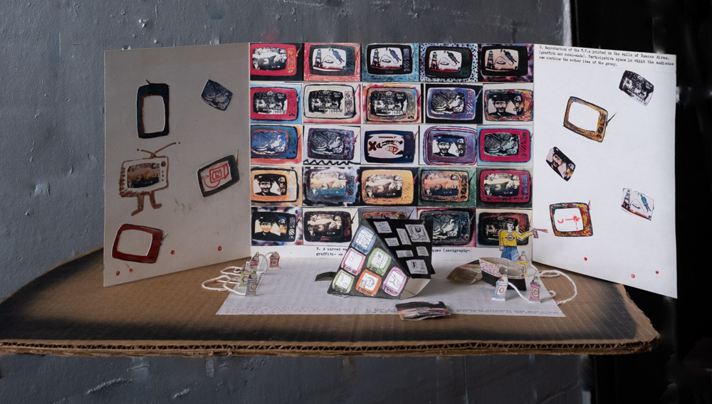 A photo of a cardboard model for the making of an exhibition. There are three "walls" made out of white cardboard standing up vertically on a piece of brown cardboard. Miniature rectangles represent artworks that are all similar in vibrant jewel tones form an edge-to-edge grid on the central panel. Other versions of the same image are scattered on the left and right panels. Mini models of sculptural elements are in the foreground on a a sheet of white paper.