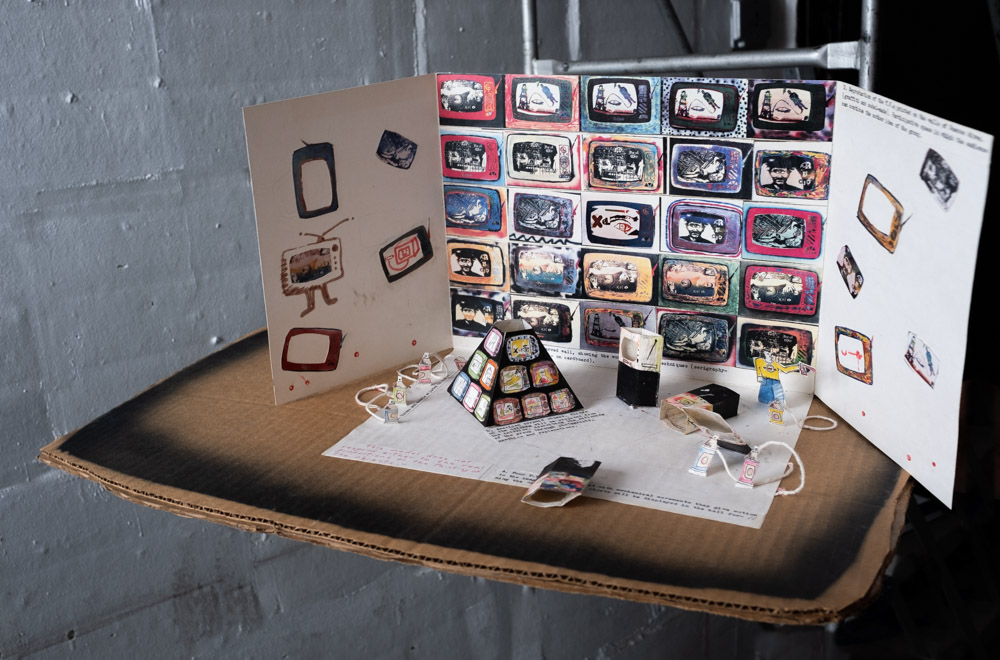 A photo of a cardboard model for the making of an exhibition. There are three "walls" made out of white cardboard standing up vertically on a piece of brown cardboard. Miniature rectangles that may represent artworks that are all similar in vibrant jewel tones form an edge-to-edge grid on the central panel. Other versions of the same image are scattered on the left and right panels. Mini models of sculptural elements are in the foreground on a a sheet of white paper.