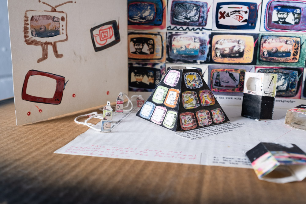 A photo of a cardboard model for the making of an exhibition. Two of the "walls" made out of white cardboard are visible. They stand up vertically on a piece of brown cardboard. Miniature rectangles that may represent artworks that are all similar in vibrant jewel tones form an edge-to-edge grid on the right hand panel. Other versions of the same image are scattered on the left panels. Mini models of sculptural elements are in the foreground on a a sheet of white paper.