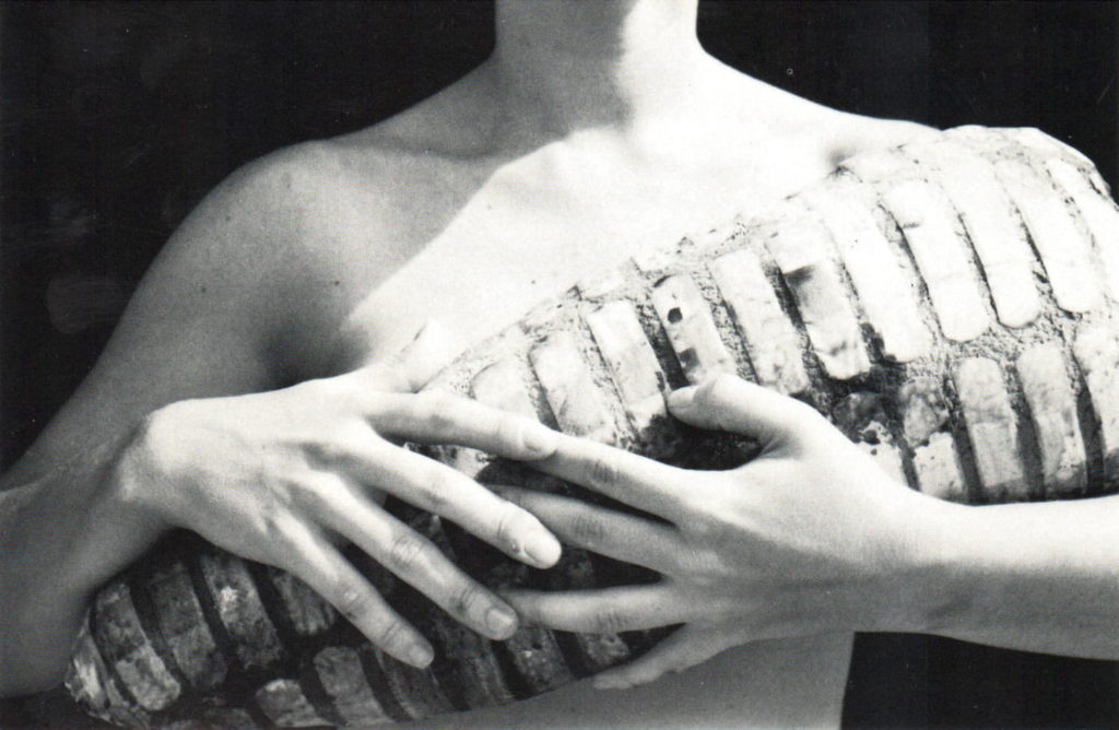 A black-and-white photo of a person from the top of their neck to mid-torso. Their arms cradle a piece of masonry across their chest. The background is dense black.