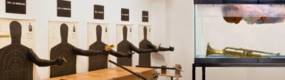 A photograph of an art installation, at left three-dimensional arms have been added to five shooting targets afixed to a wall. The right side of the image comprises a large fish tank filled with water with a gold trumpet or trombone at the bottom and a person (or mannequin) floating above, face down.