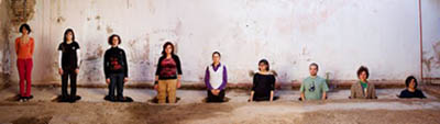 A photograph of nine people wearing every-day clothing they stand on a dirt floor with a white wall behind them. The person on the left stands on the ground, and each person to the right of them stands, incrementally, in a deeper hole in the floor. The person on the right edge of the photo is only visible from the shoulders up above the soil.