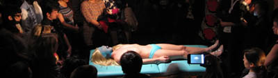 A photo of a performance where a fair-skinned person wearing turquoise brief underwear lies on a turquoise table surface. They have a blue covering over their face, and their long blonde hair rests on the table around their head like a halo. There is a light on them, and the audience surrounding the person is in darkness but several people are visible.