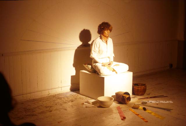 A photograph of a performance featuring a person wearing all white, sitting cross-legged on a white cube. In front of them are symmetrically arranged objects and baskets on the floor and a faint drawing is visible on the wall behind them.