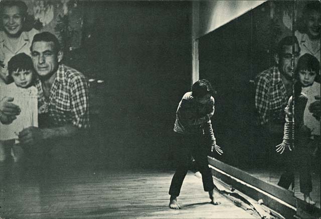 A black-and-white performance still photo of a person in a constrained pose standing between two large blown up photos of a family.