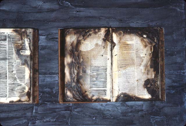 A color photograph of an open book whose pages have been burned and charred. Some of the surviving text appears to be redacted.