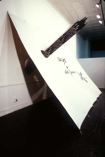 An installation photo of an artwork where a white wall is the support for a piece of taught fabric that is secured to the floor several feel away, making a tent-like shape. A skyscraper-like shape in black pierces the fabric through a hole. There is some hand written black text on the white tarp as well.