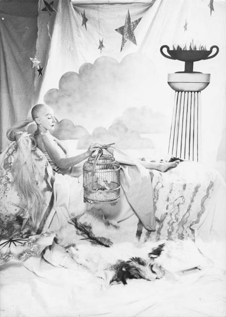 A black-and-white drawing of a bald person lounging on a chaise with a bird cage in the foreground, and a column supporting a large chalice holding flames. A large piece of fabric with clouds and stars completes the background.
