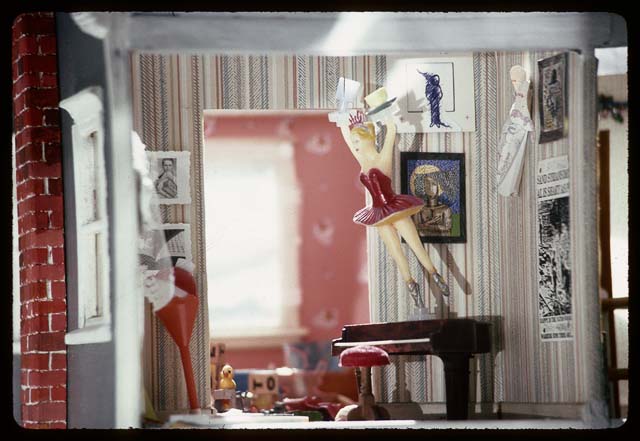 A photograph of a miniature interior scene of an apartment, like a view into a doll's house. A toy ballerina twirls above a toy piano.