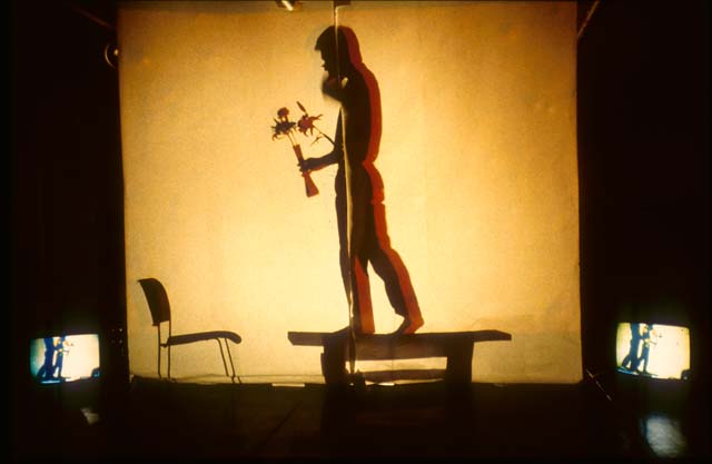 A photograph of a performance in which a yellow light silhouettes a darkened figure in profile standing on a platform holding a slim vase of flowers. A chair can be seen in front of them as they face the left side of the image. Two video monitors flank the scene.