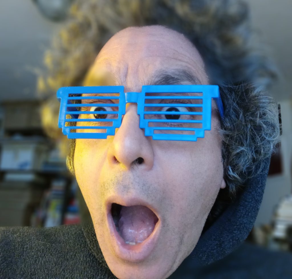 Nicolás, Middle-age person of mixed-race background with mouth wide open as if surprised pleasantly by an unexpected occurrence. Longish wavy hair sticking up. He is wearing plastic blue spectacles with horizontal stripes across where glass would usually be.