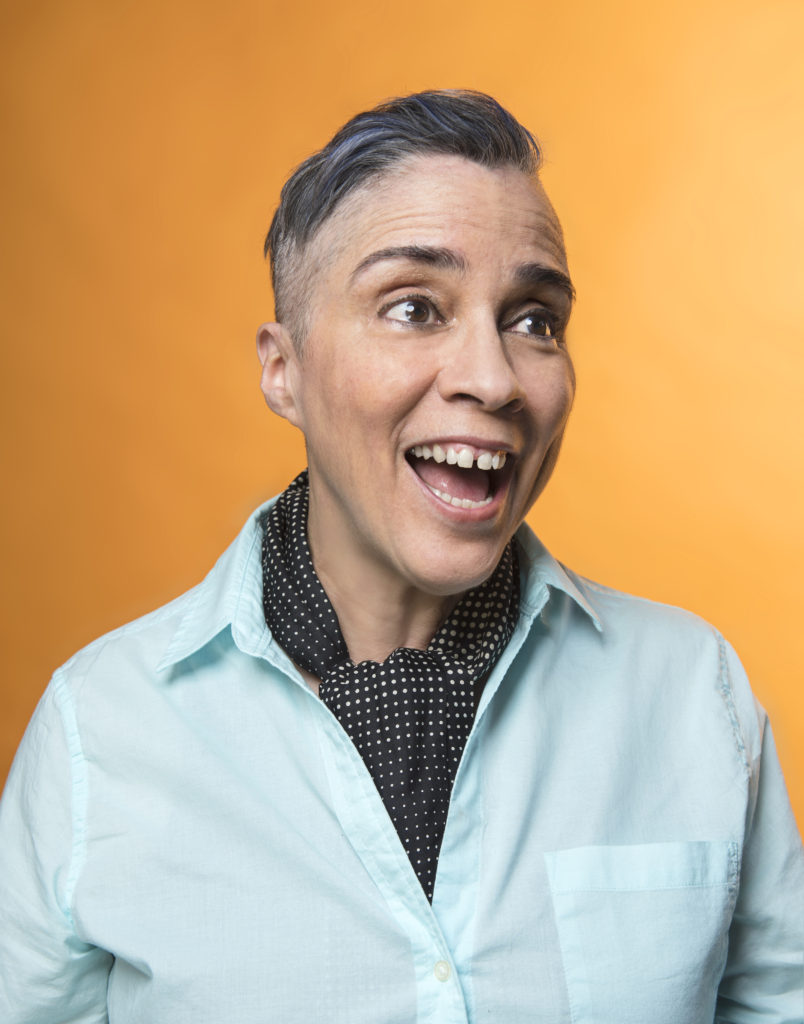 Medium photo of salt and pepper short haired gender non-conformist smiling off to the right, with a big gap toothed smile. Person is in front of an orange background wearing a light blue cotton shirt and a black and white polka dot ascot.