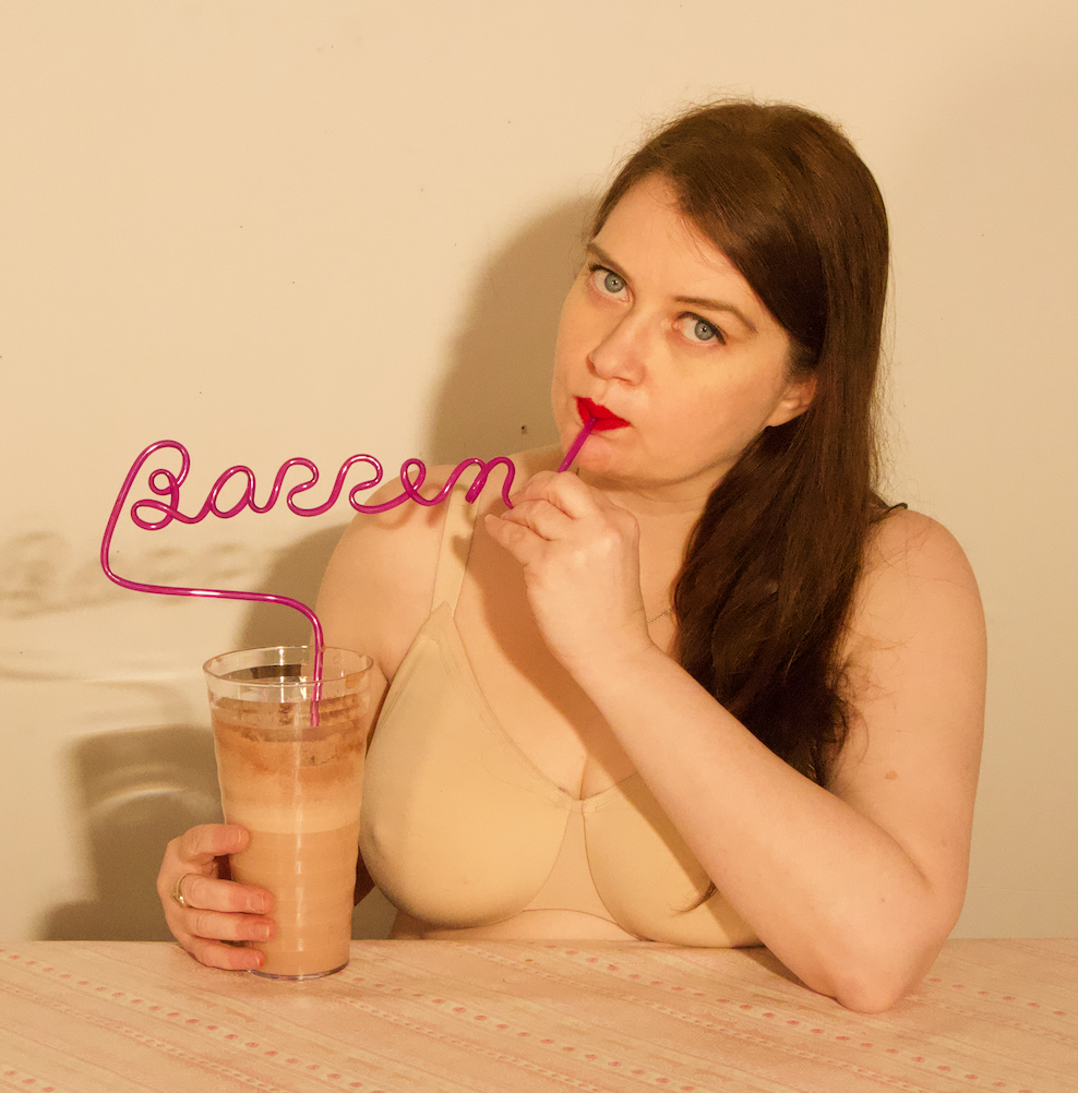 A woman with long brown hair in a nude color bra drinks a chocolate milkshake out of a straw that is molded to say the word “barren” as she looks into the camera