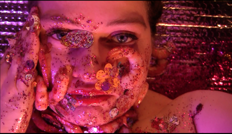 A closeup of a woman in a pink and silver background looking at the camera. Her face is covered with shrimp and glitter.